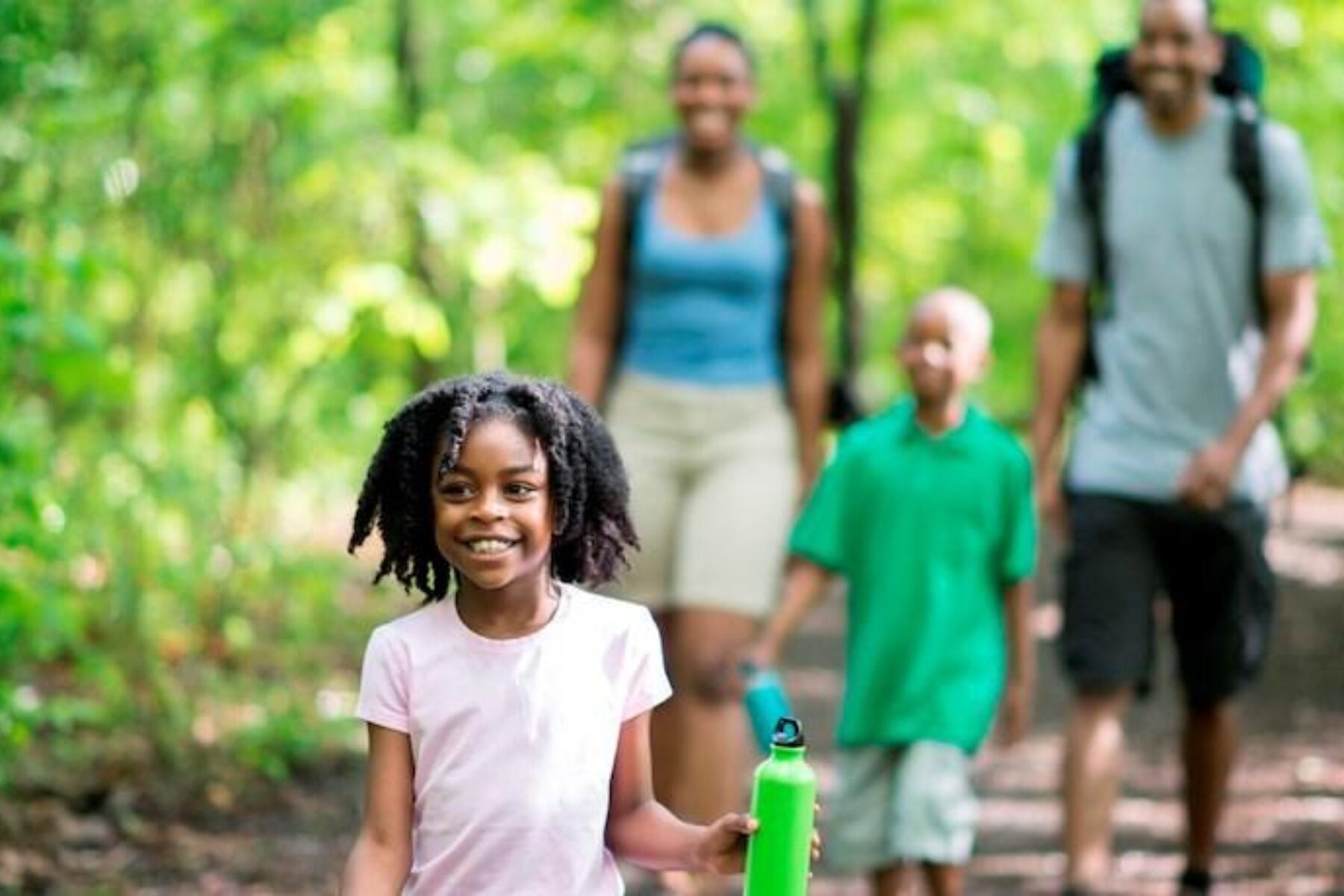 A family of four are hiking through the woods on a beautiful sunny spring day. The are walking on a footpath and there are green trees all around them. (A family of four are hiking through the woods on a beautiful sunny spring day. The are walking on