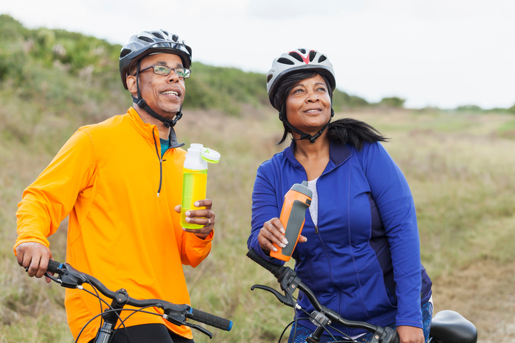 A mature, African American couple riding mountain bikes in a state park, wearing helmets. They are taking a break, drinking from water bottles, smiling and looking up.